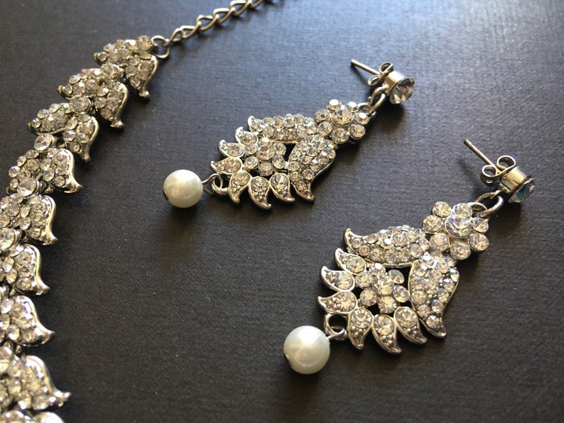 Op de afbeelding: Een zilverkleurige sieradenset bestaande uit een ketting en twee oorbellen. Elk sieraad heeft een bladvormig ontwerp ingelegd met heldere strass-steentjes en een enkele witte kunstparel die aan de onderkant van de oorbelhangers bungelt.