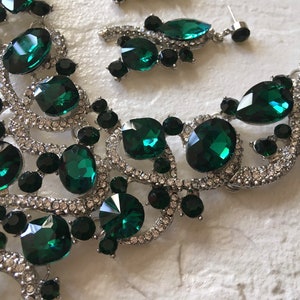 May include: A silver-toned necklace and earring set featuring large, faceted emerald green crystals, smaller round black stones, and clear pav&eacute; rhinestones set in a swirling, intricate metal frame that connects the various gemstone shapes.