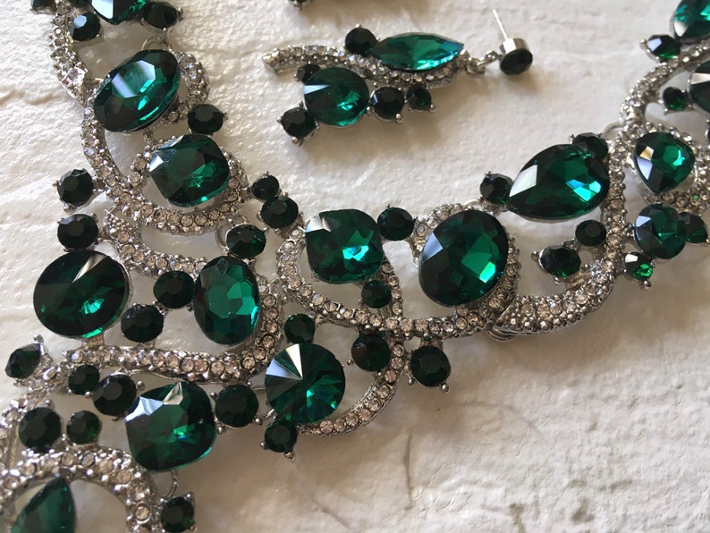 May include: A silver-toned necklace and earring set featuring large, faceted emerald green crystals, smaller round black stones, and clear pav&eacute; rhinestones set in a swirling, intricate metal frame that connects the various gemstone shapes.