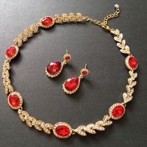 May include: A gold necklace with red gemstone accents and matching earrings. The necklace features a leaf design with clear rhinestones. The earrings are teardrop shaped with red gemstones.