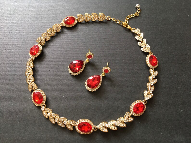 May include: A gold necklace with red gemstone accents and matching earrings. The necklace features a leaf design with clear rhinestones. The earrings are teardrop shaped with red gemstones.