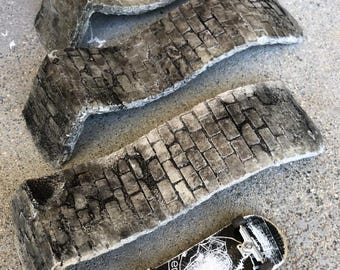 May include: Three concrete ramps with a brick pattern, designed for skateboarding. The ramps are arranged in a staggered formation, with the largest ramp at the bottom. A small black and white skateboard with the word "elemental" printed on it is in the foreground.