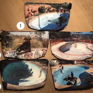 May include: Five zippered cosmetic bags with different images of skate parks and skaters. The bags are rectangular with black zippers and pulls. The images include a skater in action, a pool, and people skating in a pool.