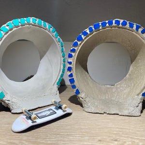 May include: Two decorative, off-white, circular planters with mosaic-style trim in turquoise and blue. A small, white skateboard with a graphic is in front of the planters. The planters have a rough, textured appearance.