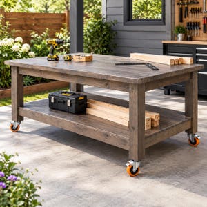 May include: A large, rectangular wooden workbench with a dark gray finish, featuring four heavy-duty swivel casters with bright orange wheels. The table includes a lower storage shelf, a yellow and black cordless drill, a black toolbox, and lumber.
