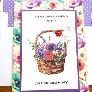 May include: A birthday card with a watercolor illustration of a basket overflowing with purple, pink, and white flowers. The card reads "To My Dear Friend Jackie on Her Birthday." The card is surrounded by floral patterned paper and a purple envelope.