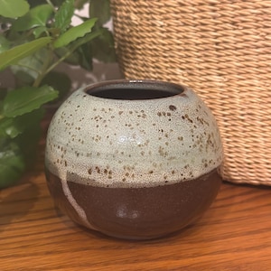 May include: A spherical ceramic vase featuring a two-tone glaze. The upper portion has a speckled light mint green finish, while the lower section is a solid dark brown. A single vertical drip of the light glaze extends down the dark base.