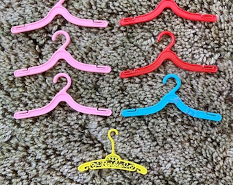 Peut inclure: Un ensemble de sept cintres miniatures en plastique de différentes couleurs. Il y a trois cintres rose clair, deux cintres rouges, un cintre bleu vif et un cintre jaune portant l'inscription Barbie en écriture cursive.