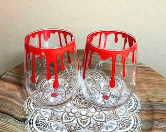 Può includere: Due bicchieri da vino in vetro trasparente con vernice rossa che cola intorno al bordo. I bicchieri sono su un pizzo bianco su una superficie di legno.