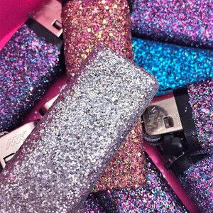 May include: A collection of lighters decorated with glitter in various colors, including silver, pink, blue, and purple. The lighters are arranged in a pile, with some overlapping.