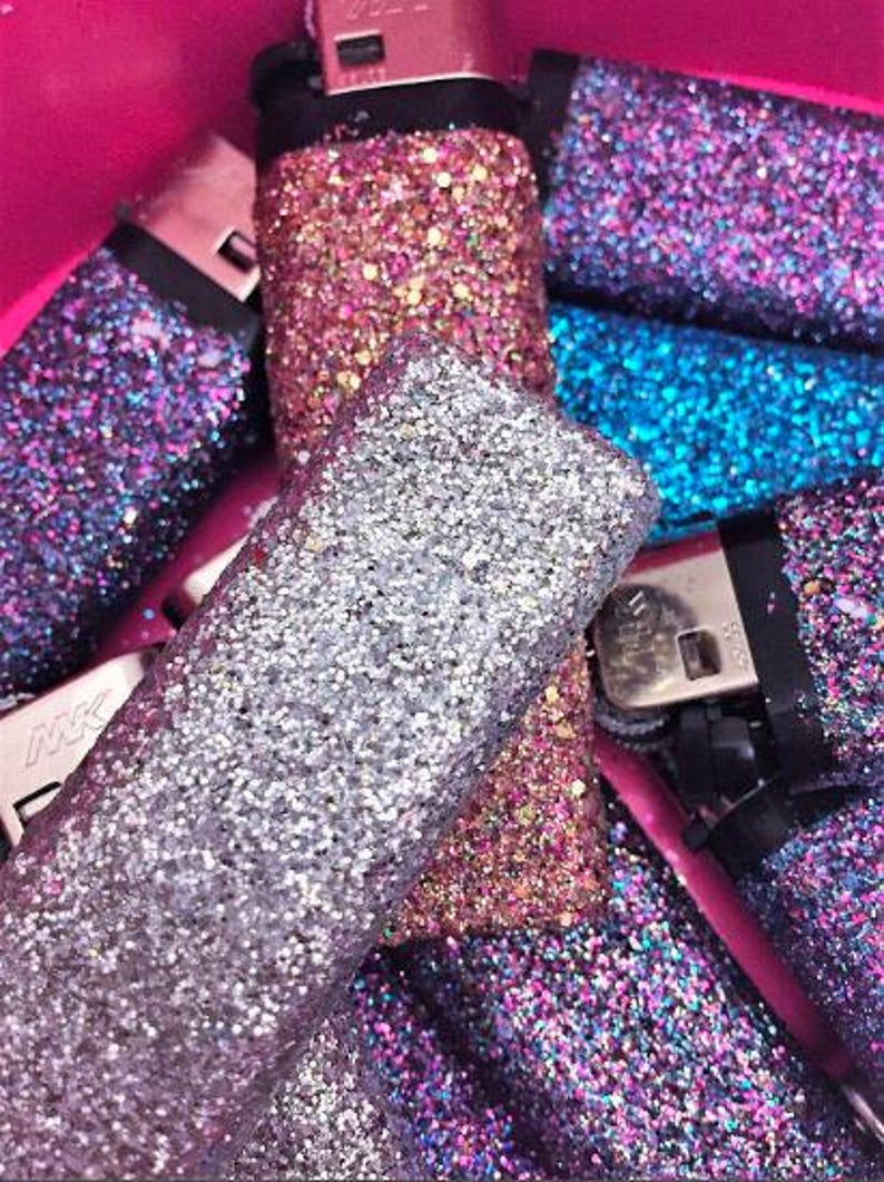 May include: A collection of lighters decorated with glitter in various colors, including silver, pink, blue, and purple. The lighters are arranged in a pile, with some overlapping.