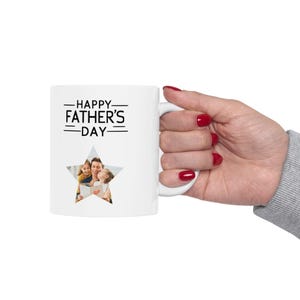 May include: White ceramic mug with the text "HAPPY FATHER'S DAY" in black. A star-shaped cutout reveals a family photo. The mug is held by a hand with red nail polish.