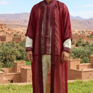 Peut inclure: Une robe transparente bordeaux portée sur une tunique et un pantalon assortis de couleur crème. La robe présente une broderie foncée complexe le long de l'ouverture avant et des poignets, avec un col montant et une bordure décorative.