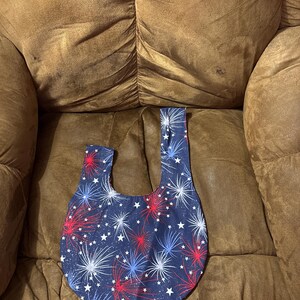 Könnte beinhalten: Eine Stofftasche mit Handgelenkschlaufe auf dunkelblauem Grund mit einem sich wiederholenden Muster aus roten, weißen und blauen Feuerwerken sowie kleinen weißen Sternen. Die Tasche hat ein asymmetrisches Griffdesign mit einer längeren Seite.