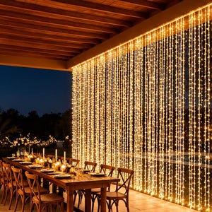 Peut inclure: Un rideau de guirlandes lumineuses LED blanc chaud suspendu verticalement à un plafond de terrasse en bois. Les lumières créent une toile de fond dense et lumineuse derrière une longue table à manger en bois dressée sur une terrasse en pierre.