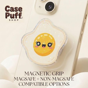 May include: A phone grip featuring a glittery, fried egg design with a smiling face, attached to a light-colored smartphone. The text Case Puff SHOP, MAGNETIC GRIP, MAGSAFE + NON-MAGSAFE COMPATIBLE OPTIONS is visible on the image.