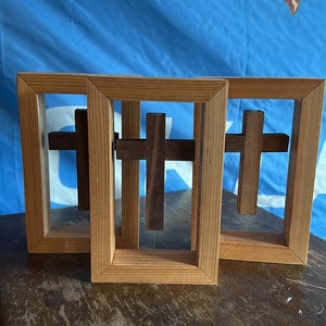 May include: Three wooden frames, each containing a dark brown wooden cross suspended in the center. The frames are made of light-colored wood with a natural grain, and the crosses appear to be floating within the rectangular openings.