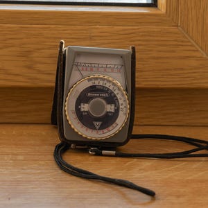 May include: A vintage grey light meter with a circular dial, white and blue face, and the text Leningrad printed in the center. The device features a black leather case and a thin black neck strap, displaying various numerical scales and settings.