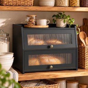 May include: A black two-tier bread box featuring fluted glass panels on the front of each drop-down door. Each door is equipped with a small, round, metallic knob. The structure is rectangular and provides storage for baked goods on kitchen shelves.