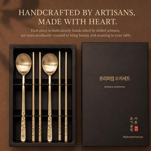 May include: A set of two gold-toned metal spoons and two pairs of matching chopsticks in a black presentation box. The handles feature engraved floral patterns. The box lid displays the text: HANDCRAFTED BY ARTISANS, MADE WITH HEART, 프리미엄 수저세트, SPOON & CHOPSTICKS, and MyKoreanFortune.