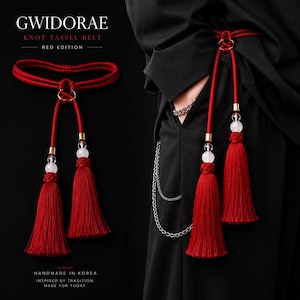 May include: A red knot tassel belt labelled GWIDORAE KNOT TASSEL BELT RED EDITION. The belt features a circular gold-tone ring, two long red cords, and two large red tassels accented with clear spherical beads and gold-tone metal hardware.