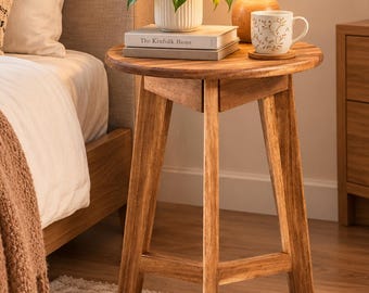 May include: A round wooden side table with three angled legs. On the table sits a white ribbed pot with a green plant, a book titled The Kinfolk Home, a white lamp with a wooden base, and a white mug with a gold leaf pattern on a cork coaster.