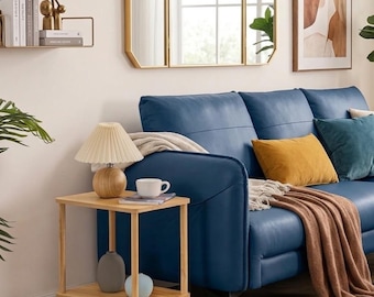 May include: A two-tiered light wood side table featuring a round lamp with a pleated shade, a white mug, and two small vases on the lower shelf. The table is positioned next to a blue sofa with mustard and teal pillows and a brown throw blanket.