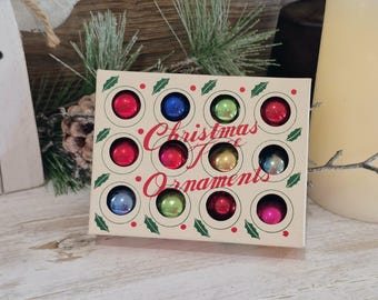 May include: A rectangular cream-colored box containing twelve small, round, multi-colored Christmas ornaments. The box features the text "Christmas Ornaments" in red script, surrounded by green holly leaf illustrations and small red dots.