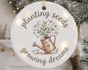 May include: A round white ceramic ornament featuring a gold watering can with a heart icon, a green plant sprouting from the top, and the text "planting seeds growing dreams" in dark green script, suspended by a gold string.