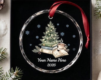 May include: A round, clear glass ornament with a faceted edge, featuring a printed illustration of a Christmas tree, a stack of books, and a coffee cup. The text reads Your Name Here and 2026. A red ribbon is attached for hanging.