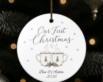 May include: A round white ceramic ornament featuring the text Our First Christmas, Ben & Ashlee, and 2026. The design includes two steaming mugs with small pine tree illustrations, a heart, and light blue snowflakes on a white background.