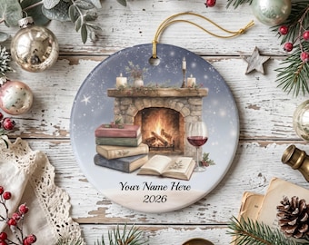 May include: A round ceramic ornament featuring a fireplace illustration with a stack of books, an open book, and a glass of red wine. The text "Your Name Here" and "2026" is printed at the bottom. The ornament has a gold hanging cord.