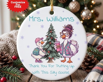 May include: A round white ceramic ornament featuring a cartoon illustration of two geese near a Christmas tree. The text reads Mrs. Williams and Thank You For Putting Up With This Silly Goose. The ornament hangs from a gold string.