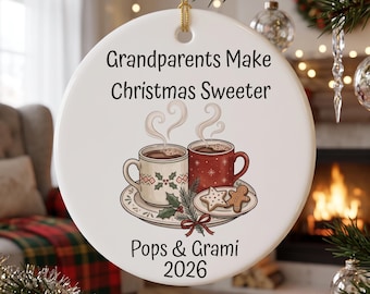 May include: A round white ceramic Christmas ornament featuring the text Grandparents Make Christmas Sweeter, Pops & Grami, and 2026. The design includes two mugs of hot cocoa, a plate with a star cookie and gingerbread man, and holly.