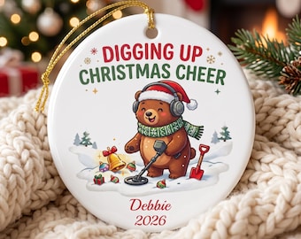 May include: A round white ceramic ornament with a gold hanging string. The design features a brown bear wearing a Santa hat, headphones, and a green scarf, using a metal detector in the snow. Text reads DIGGING UP CHRISTMAS CHEER, Debbie, 2026.