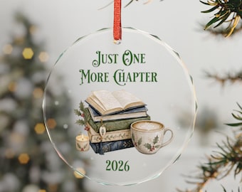 May include: A circular glass ornament hanging from a red ribbon, featuring the text "Just One More Chapter" and "2026" in green. The design includes a stack of four books, a small lit candle, and a mug with a latte art design.