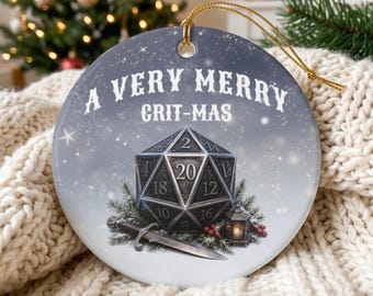 May include: A round ceramic ornament with a gold string, featuring a gray background with white text that reads A VERY MERRY GRIT-MAS. The design includes a twenty-sided die, a silver sword, a small lantern, and pine branches with red berries.