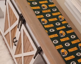 May include: A wooden console table with a white frame and natural wood panels, featuring sliding barn-style doors with black metal hardware. The top surface is covered in a green fabric runner printed with the Green Bay Packers logo and text.
