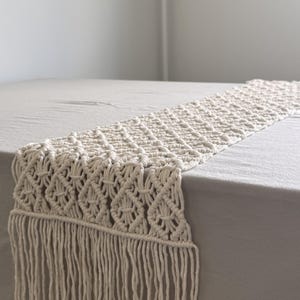 May include: An off-white macrame table runner featuring a repeating diamond knot pattern and long, straight fringe along the edge. The textile is crafted from thick cord and displays a textured, woven design that extends across the surface.