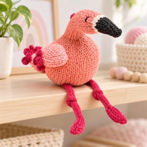 May include: A handcrafted knit flamingo plush toy featuring a round coral-pink body, light pink wings with dark pink ruffled edges, and a black beak with a white base. The stuffed animal has long, thin, dark pink legs with knotted joints.