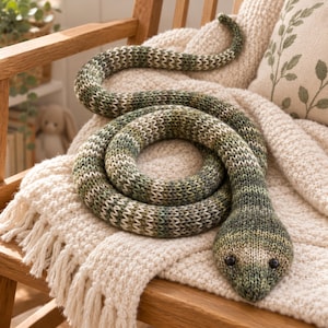 May include: A long, coiled snake plush toy crafted from variegated yarn in shades of forest green, olive, and cream. The stuffed animal features a textured knit pattern throughout its body and two small, round black buttons serving as eyes.
