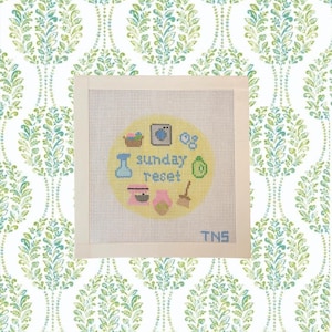 May include: A square needlepoint canvas featuring a yellow circle with the text "sunday reset" in blue. Icons include a laundry basket, washing machine, bubbles, spray bottle, green sponge, pink mixer, pink bottle, and a broom, with "TNS" in the corner.