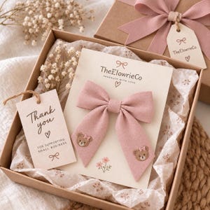 Può includere: Un fiocco per capelli in tessuto rosa polvere con due facce d'orso ricamate sulle estremità, presentato su un cartoncino con il marchio TheElowrieCo. Il fiocco è all'interno di una scatola regalo con un biglietto di ringraziamento.