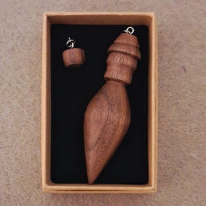 May include: A dark brown wooden pendulum with a teardrop shape and a ribbed top section, accompanied by a small matching wooden bead, both featuring silver-toned metal loops, presented inside a rectangular cardboard box with black lining.
