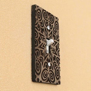 Peut inclure: Une plaque de recouvrement d'interrupteur rectangulaire en bois brun foncé avec un motif complexe découpé au laser composé de lignes tourbillonnantes et de formes géométriques. Fixée par deux vis blanches, elle entoure un interrupteur blanc.