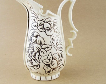 May include: Cream-colored ceramic pitcher with a black floral design. The pitcher has a curved handle and a spout. The floral pattern covers the body of the pitcher. The pitcher is on a neutral background.