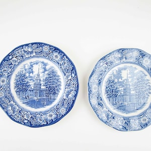 May include: Two blue and white ceramic plates with a floral border and a central image of a building with a steeple. The plates are on a white background.