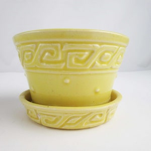 May include: A pale yellow ceramic planter with a matching saucer. The planter features a Greek key design around the top and bottom, with small raised dots. A decorative container for plants.