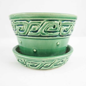 May include: A green ceramic plant pot with a matching saucer. The pot features a raised geometric Greek key pattern around the rim and a series of small raised dots around the center. The glaze is a glossy, vibrant green with darker accents.