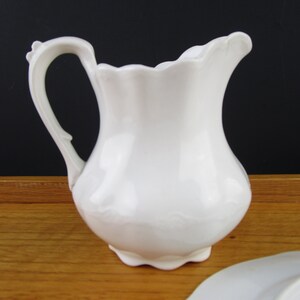 May include: A white ceramic pitcher with a curved handle and a scalloped rim. The pitcher has a bulbous body and sits on a small foot. A white plate is partially visible in the foreground. The pitcher is likely used for serving beverages.